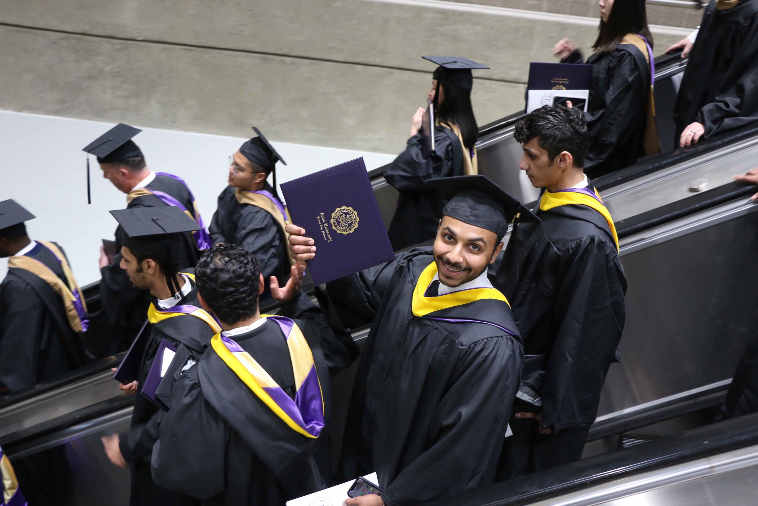 一名毕业生骄傲地拿着他的毕业证书，坐着满是应届毕业生的自动扶梯走下电梯
