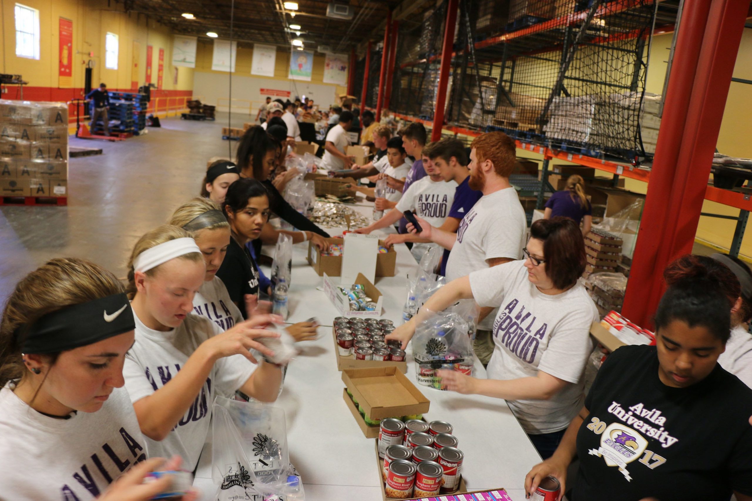 Group of students lining both sides of a long table picking and sorting canned goods inside the Harvesters warehouse