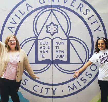 Picture of two young women posing in front of a wall with Avila University seal.