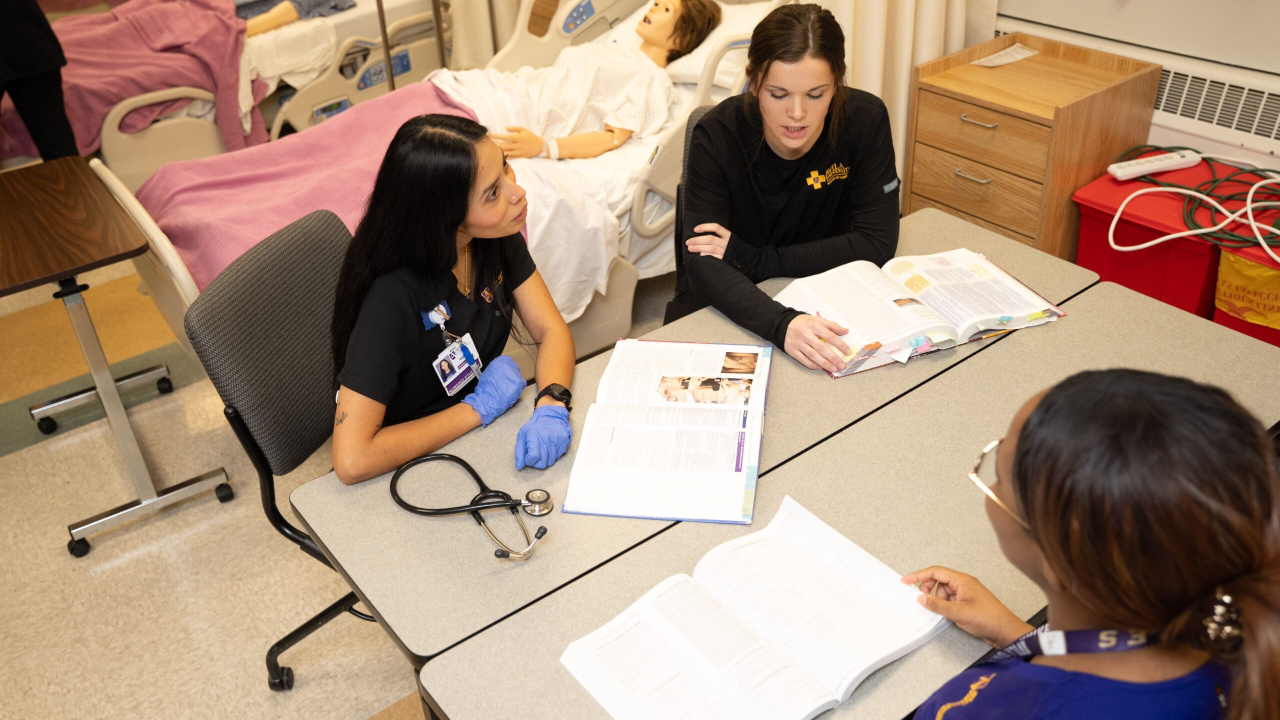 Three Avila University nursing students in black scrubs sit around tables in a nursing simulation lab, reviewing open textbooks together. A stethoscope rests on the table in front of one student wearing blue latex gloves. In the background, a full-body patient simulator mannequin lies in a hospital bed. Medical equipment and a red sharps disposal container are visible nearby.