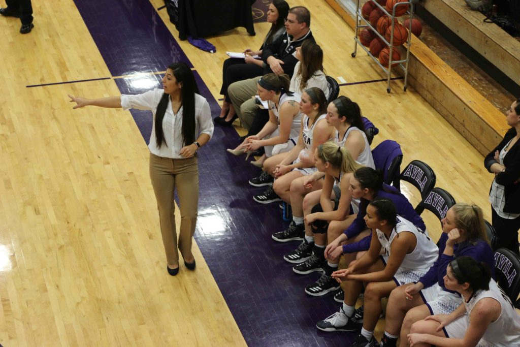 Overhead view of the Avila women's basketball team on the court side bench. Coach Murillo is calling out to players on the court