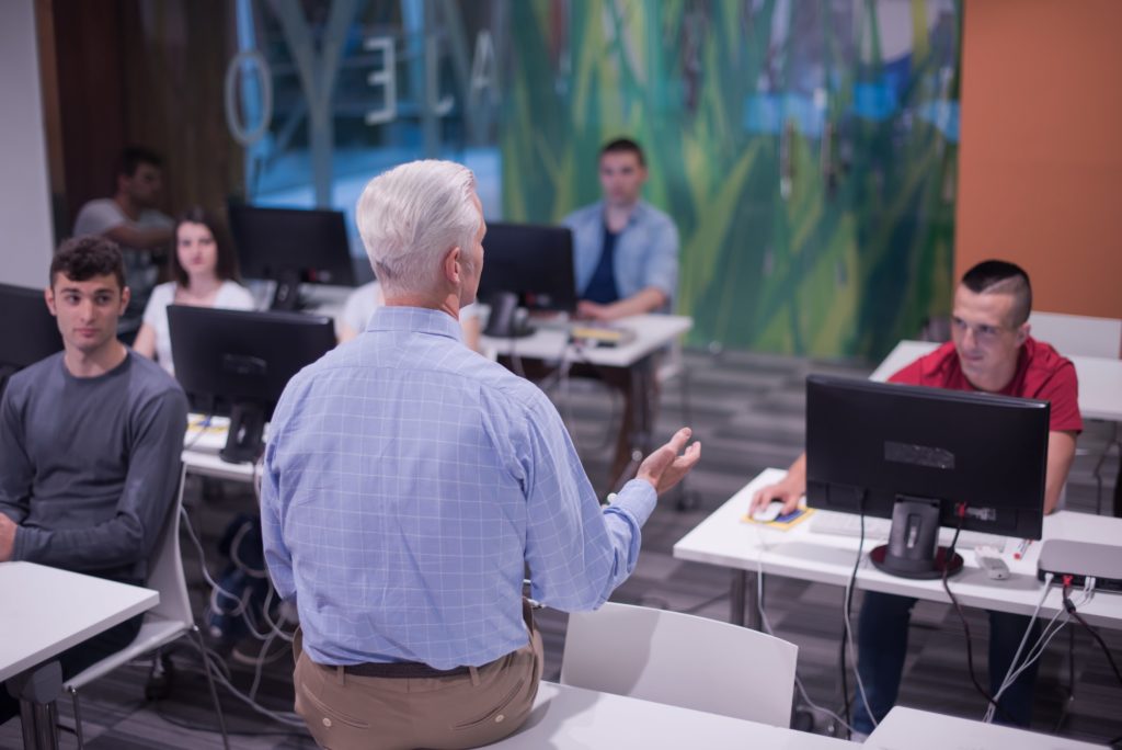 Computer science professor with back to the camera, sitting on the corner of a desk talking to three students