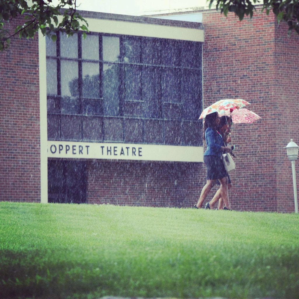 Two students holding umbrellas and walking in the rain past the Goppert Theatre entrance.