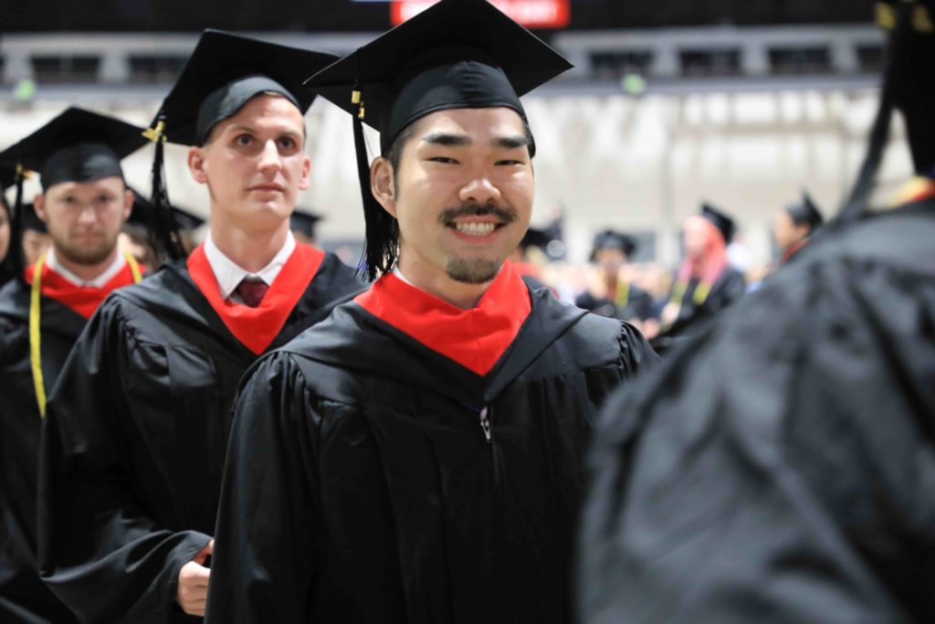 Male student smiles at camera while standing in a line of four students in caps and gowns.