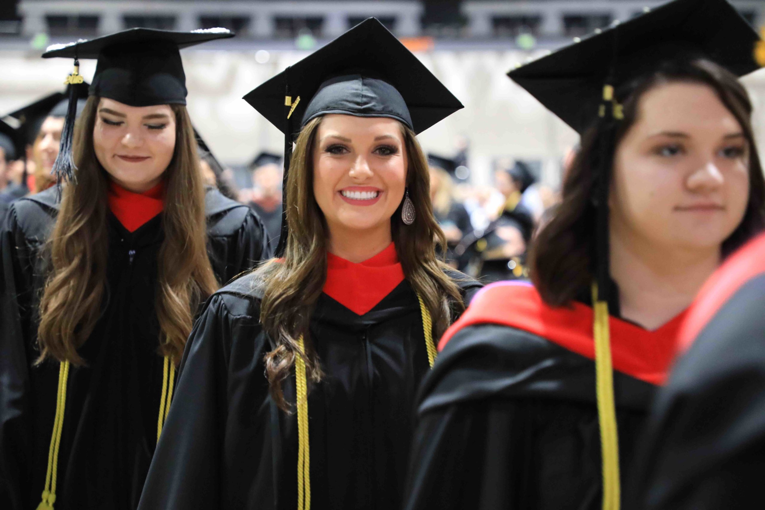 Female student smiles at the camera while standing in a line of three students wearing caps and gowns