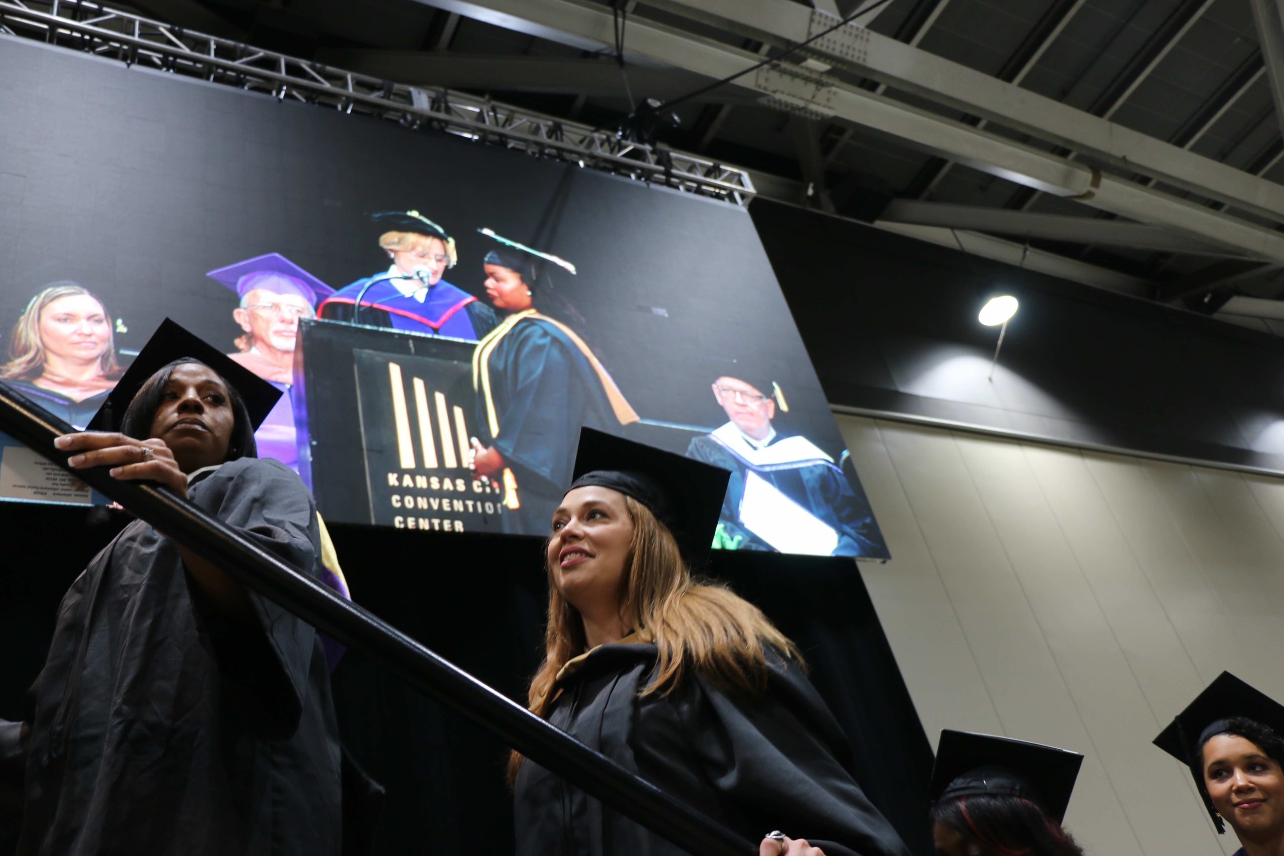 Two female students head up the stairs to the graduation stage wearing their caps and gowns