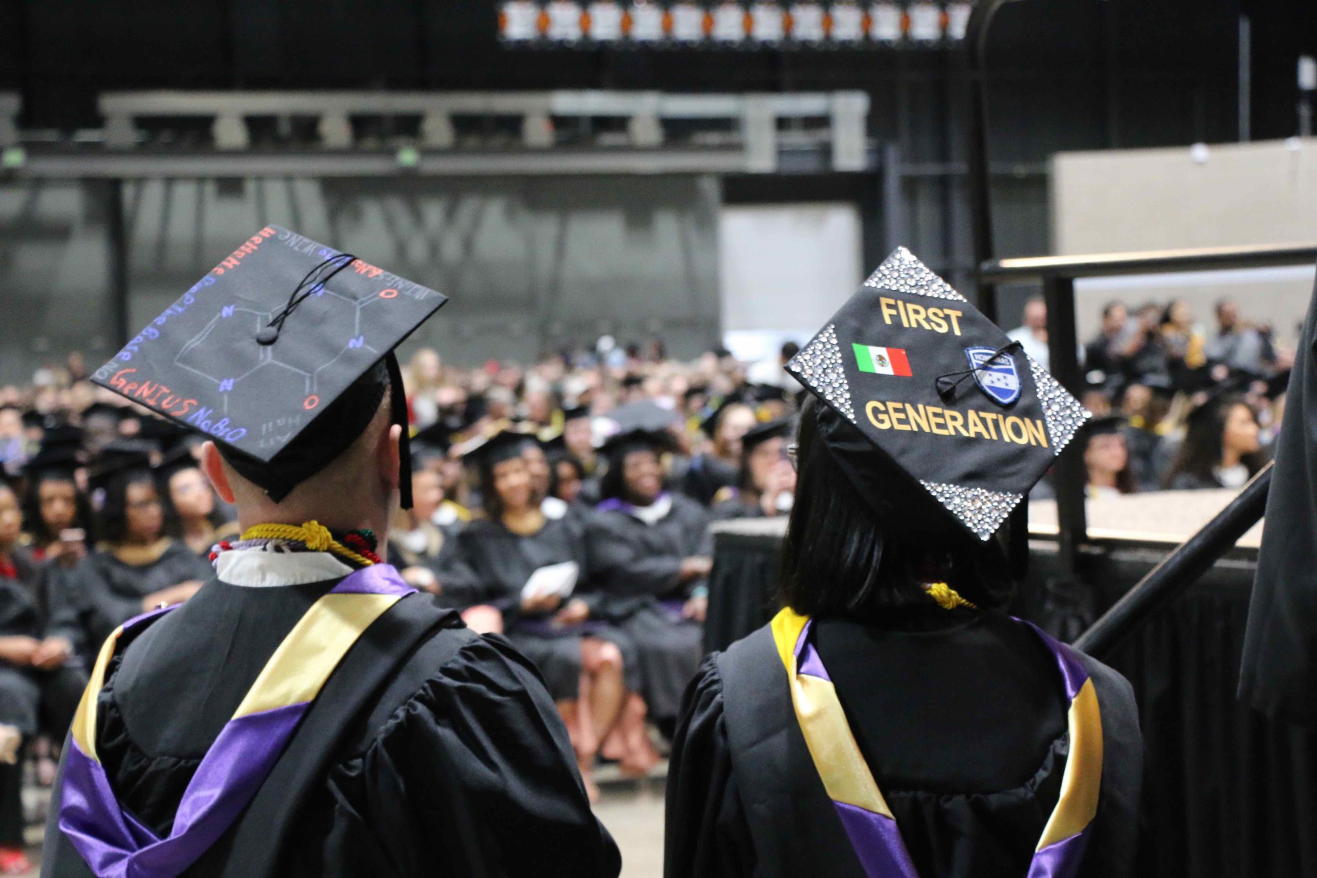 Student looking at the crowd of graduates in their caps and gowns. Her cap reads "First Generation"