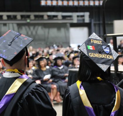 Student looking at the crowd of graduates in their caps and gowns. Her cap reads "First Generation"