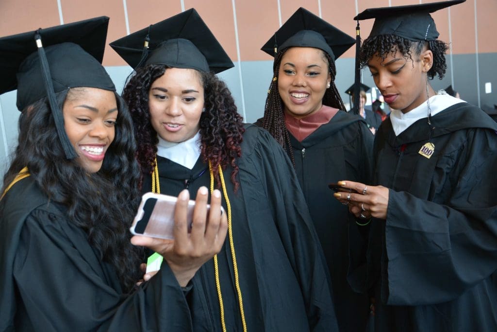 Four students stand together taking selfies in their caps and gowns