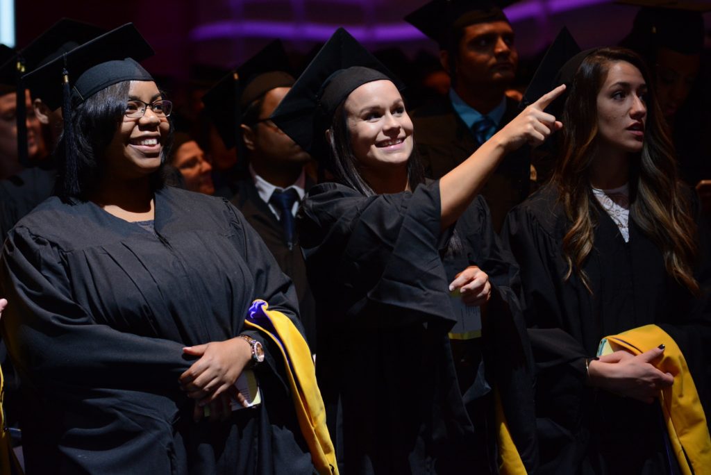 Three students point at the graduation stage while standing in the crowd wearing their caps and gowns.