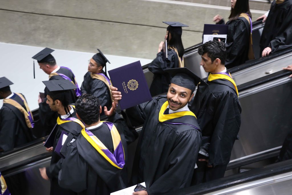 Graduate proudly holding out his diploma as he rides down an escalator filled with recent grads
