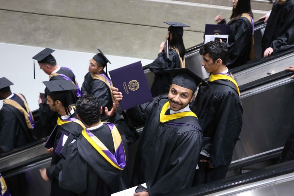 Graduate proudly holding out his diploma as he rides down an escalator filled with recent grads