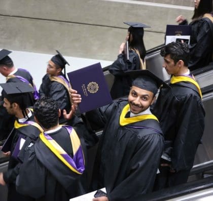 Graduate proudly holding out his diploma as he rides down an escalator filled with recent grads