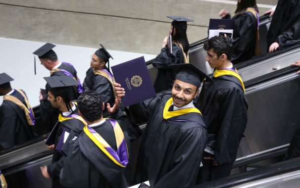 Graduate proudly holding out his diploma as he rides down an escalator filled with recent grads