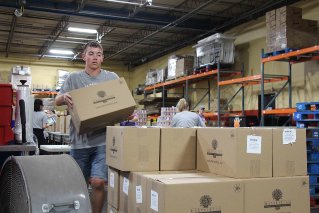 Male student inside a warehouse stacking Harvesters Community Food Network boxes on a pallet of boxes