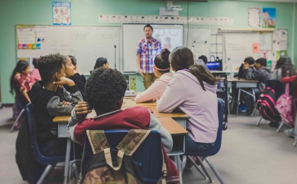 Teacher addressing a classroom of middle school students sitting at their desks.