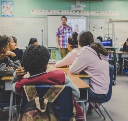 Teacher addressing a classroom of middle school students sitting at their desks.