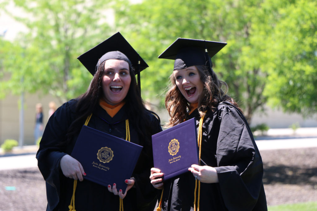 Two students in caps and gowns excitedly posing with their diplomas