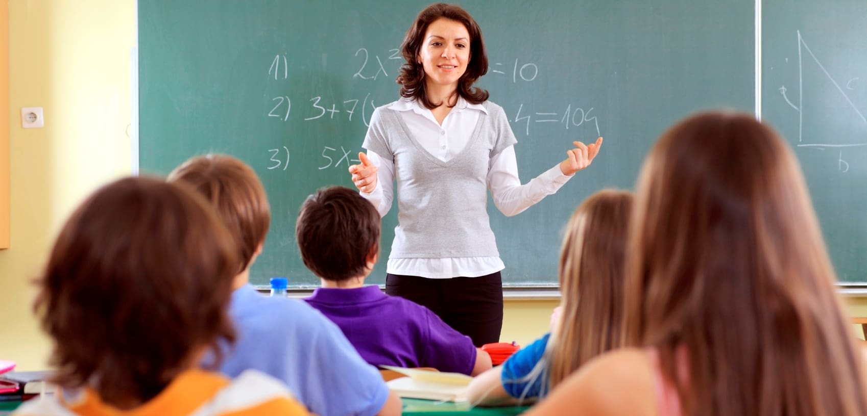 Teacher addressing a classroom of elementary students sitting at their desks.