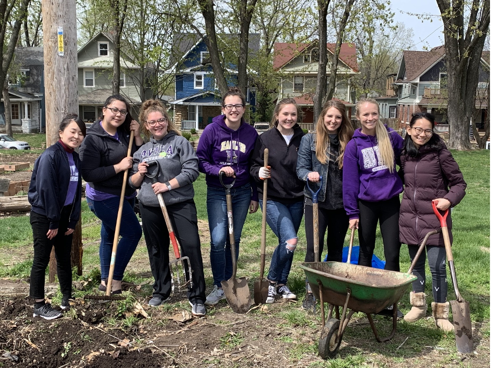 Eight students with shovels, rakes, forks, and wheelbarrow gardening at St. James