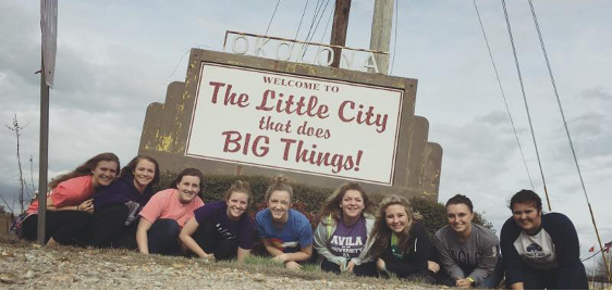 Group of students posing in front of "Welcome to the little city that does big things" sign