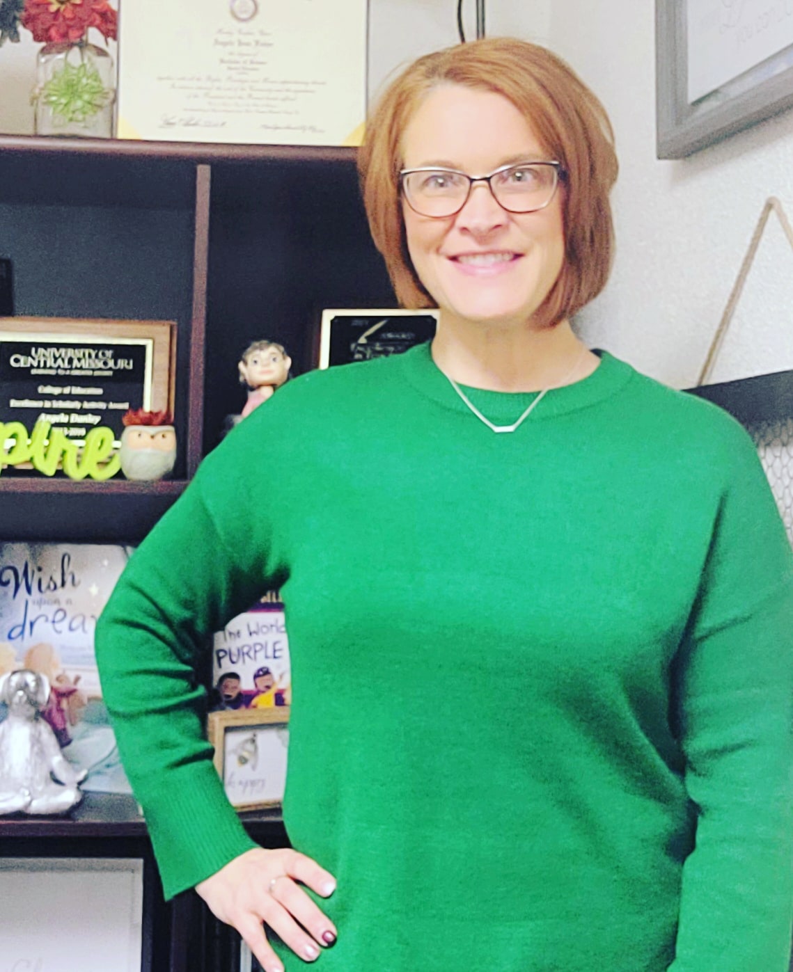 Danley is posing. She's wearing a green sweater. She has short red hair and glasses.