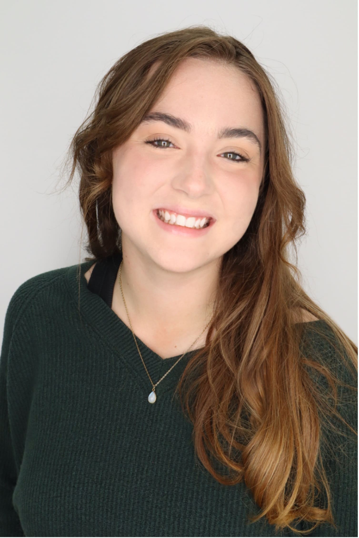 Photograph of a person with long, wavy brown hair wearing a dark green sweater and a necklace with a small pendant. The face is blurred for privacy, focusing attention on clothing and hairstyle.