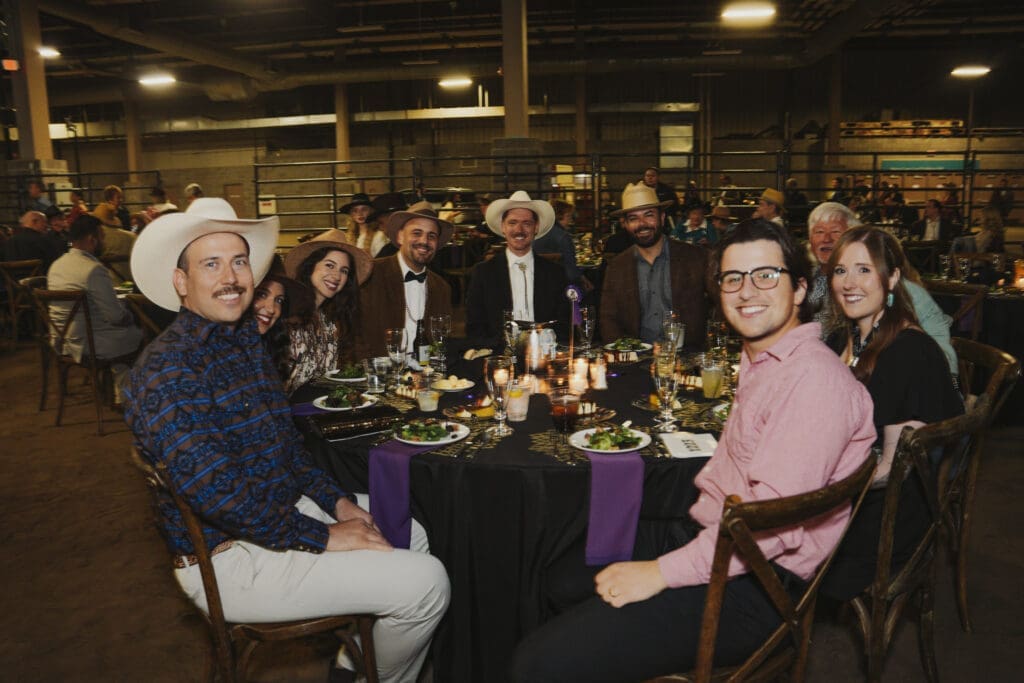 A group of guests dressed in western-themed and formal attire gather around a decorated dinner table, smiling at the camera during the Avila University 50th Steer Dinner celebration.