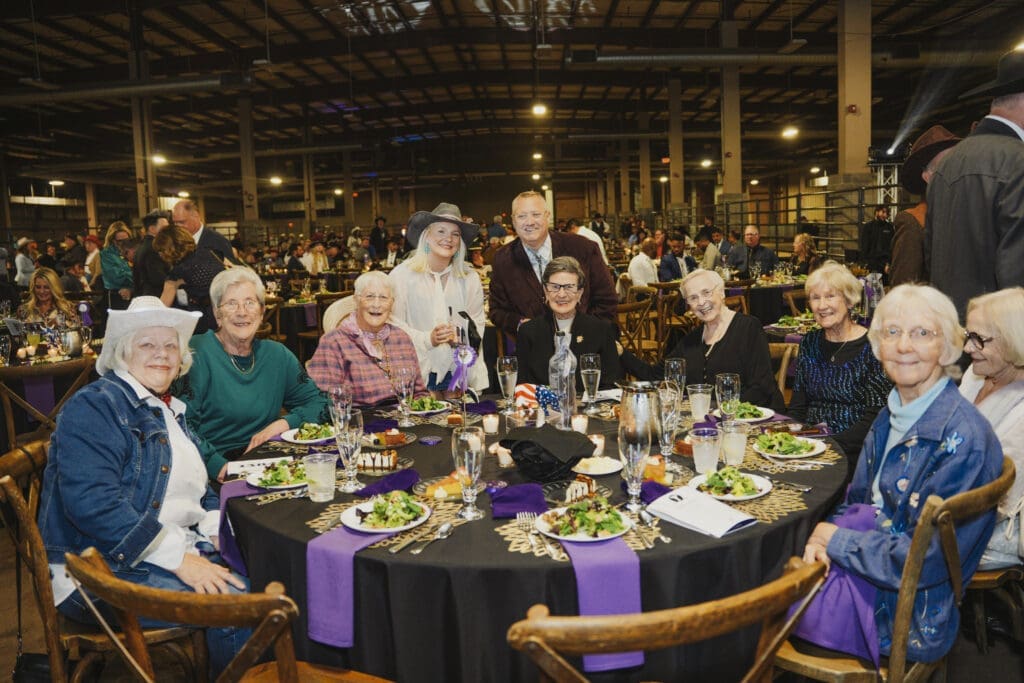 A group of Avila University supporters and guests smile together at a round dinner table elegantly set with candles, floral centerpieces, and purple linens at the 50th Steer Dinner gala.