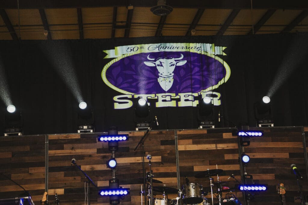 A large illuminated stage backdrop displaying the word "STEER" with a longhorn bull logo, surrounded by purple stage lighting at the Avila University 50th Steer Dinner event.
