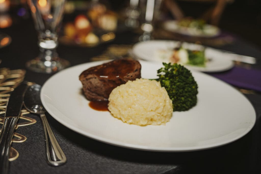 An elegantly plated dinner course featuring a protein entrée served with mashed potatoes and garnished with a sprig of herbs, presented on a white plate against a dark background.