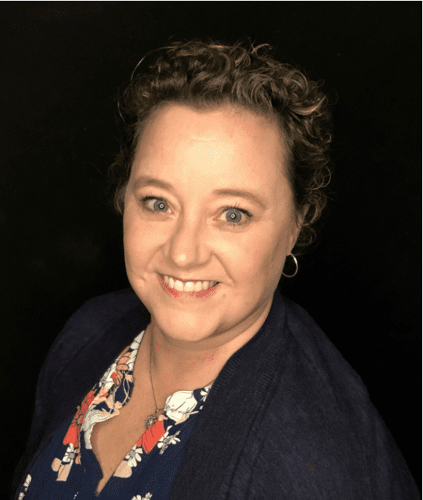 Photograph of a person with curly hair wearing a floral-patterned shirt and dark cardigan against a black background. The face is blurred to obscure identity, focusing on clothing and hairstyle details