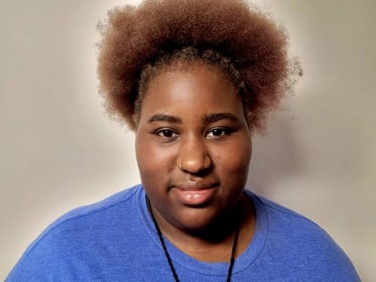 Photograph of a person with a large, curly afro hairstyle wearing a blue shirt and a black necklace against a plain, light-colored background. The focus is on the hairstyle and upper torso, with neutral lighting highlighting hair texture and clothing color.

