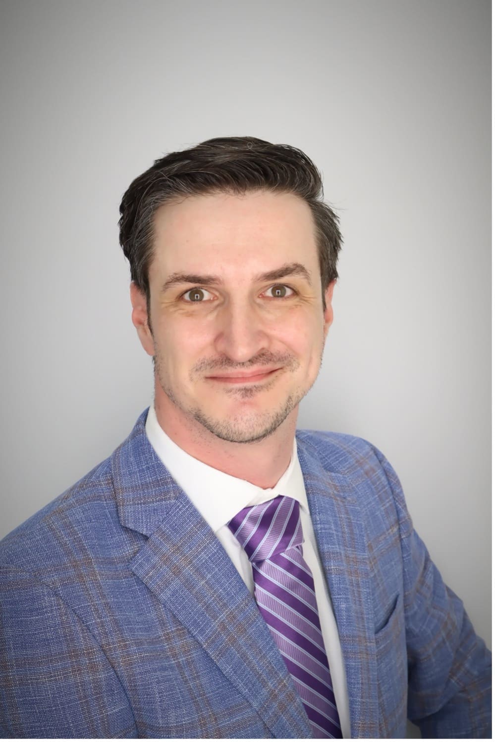 Photograph of a person wearing a blue plaid suit jacket, white shirt, and purple striped tie against a plain gray background. Face is pixelated, focusing on professional attire and hairstyle.