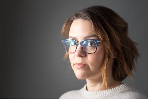 Photograph of a person with shoulder-length brown hair wearing blue-framed glasses and a light-colored sweater against a plain gray background. The face is blurred, focusing attention on hairstyle and clothing.