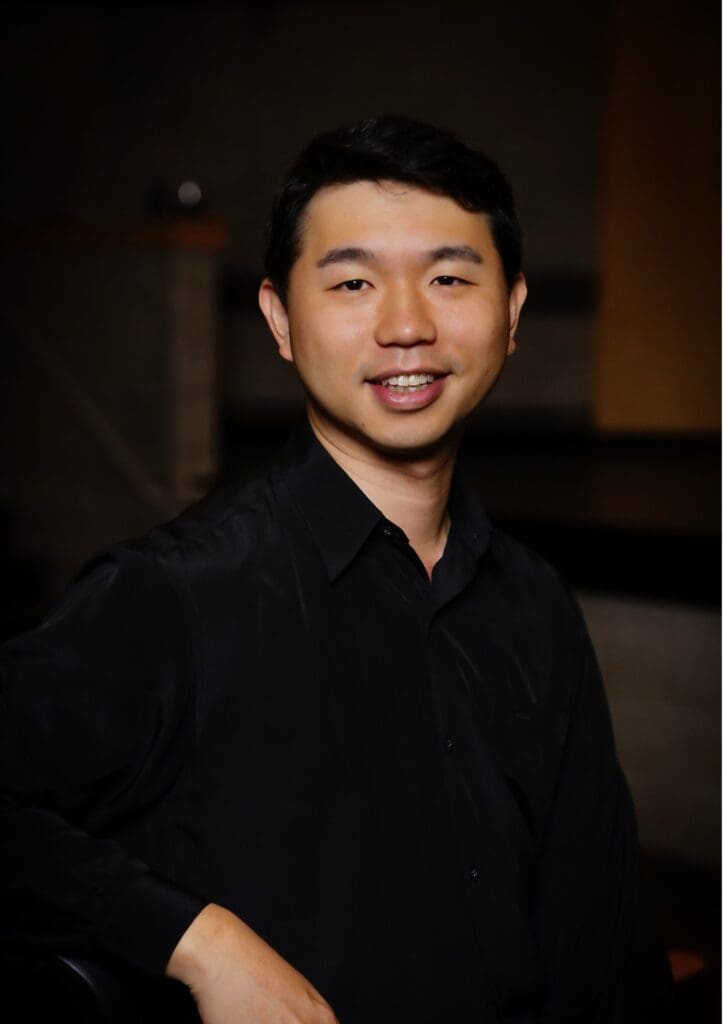 Photograph of a person wearing a black button-up shirt standing indoors with a blurred face. Background includes dim lighting and indistinct objects, suggesting a casual or professional setting.
