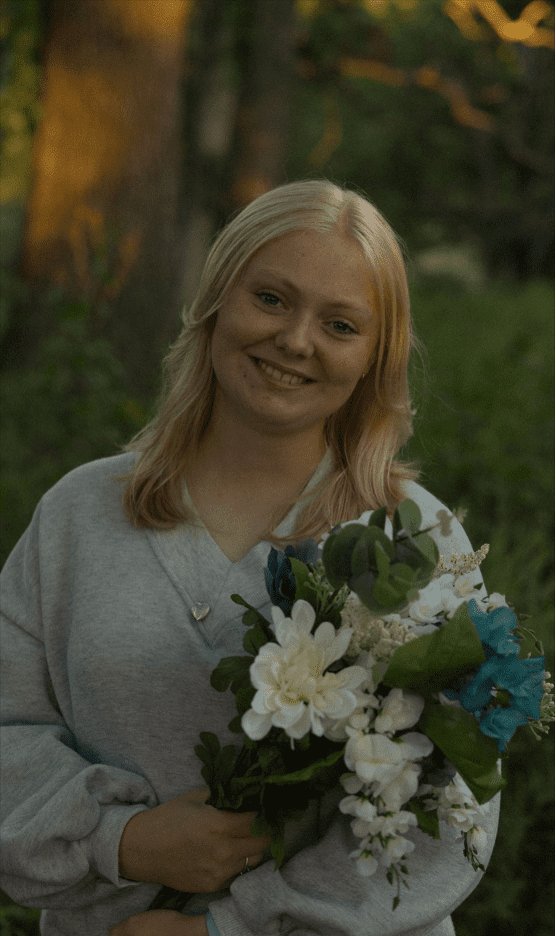 Photograph of a person with blonde hair wearing a light gray sweatshirt holding a bouquet of white and blue flowers with green leaves. Background features blurred trees and greenery, suggesting an outdoor setting during golden hour