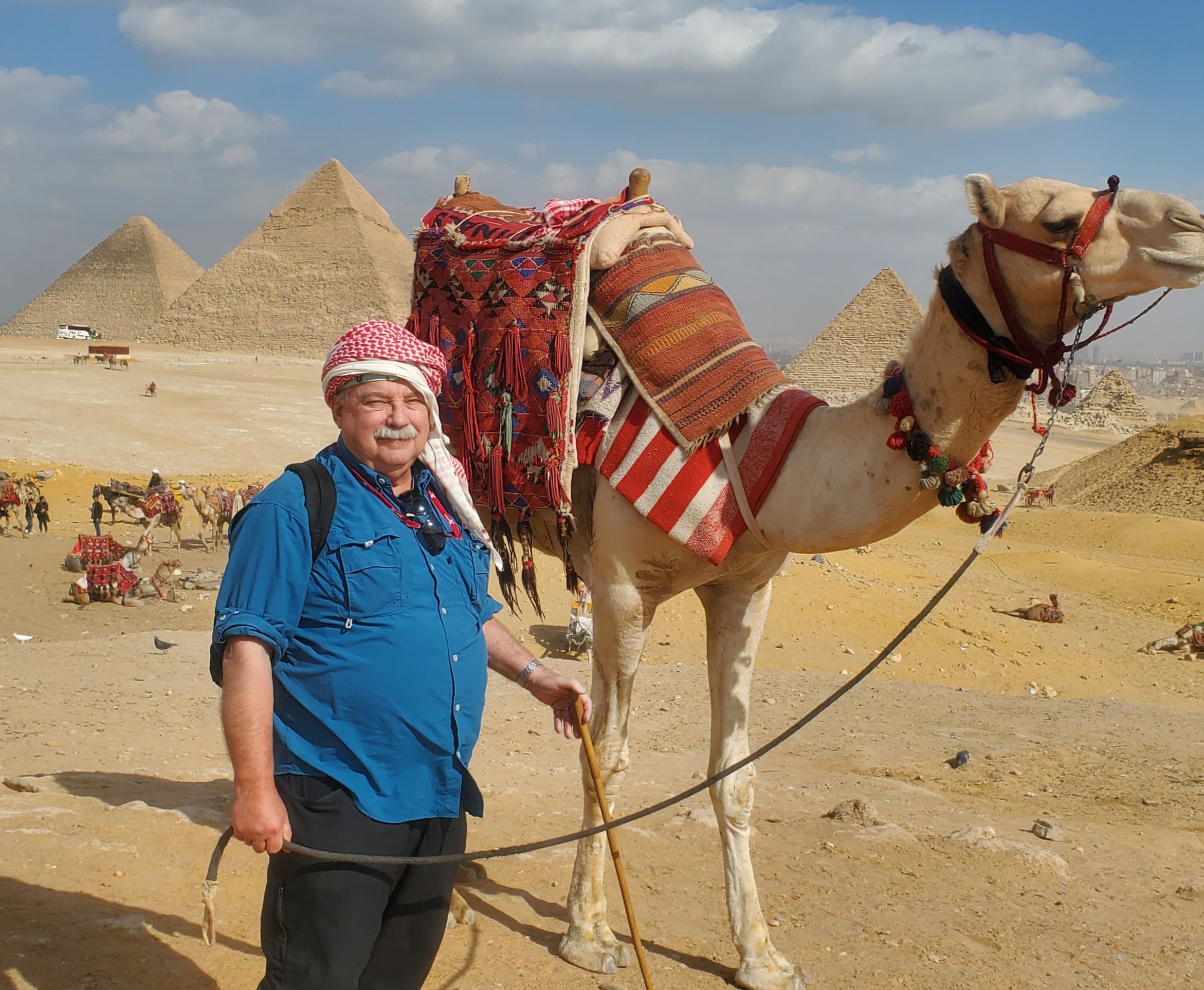 A man in a blue shirt and red-and-white keffiyeh stands holding a rope lead next to a decorated camel with colorful woven saddle blankets and tassels, with the Great Pyramids of Giza and a sandy desert landscape in the background.