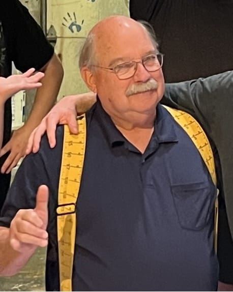 Photograph showing an older man wearing a navy blue shirt and yellow suspenders with ruler markings, giving a thumbs-up gesture. Two other people partially visible, one with an arm around the man's shoulder, set against a background with handprints on a wall.