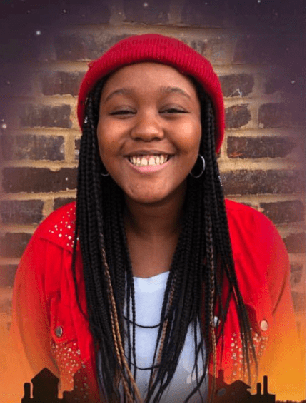Photograph of a person wearing a red beanie and red jacket with white embellishments, standing against a brick wall background. The person has long braided hair and is dressed in a white shirt underneath the jacket.