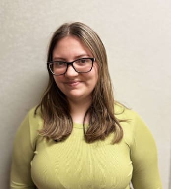 Photograph of a person with shoulder-length brown hair wearing a light green ribbed long-sleeve shirt, standing against a plain light-colored background. The person's face is blurred to obscure identity, and they are positioned facing forward.