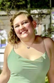 Photograph of a person wearing a light green sleeveless top with thin straps, standing outdoors at night with blurred trees and building in background. The person has curly hair and is wearing a small necklace with a pendant.