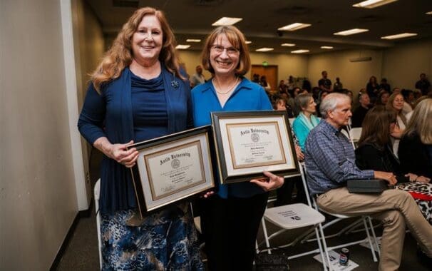 Two women smile while holding framed Avila University certificates at an award ceremony, with an audience seated in the background.