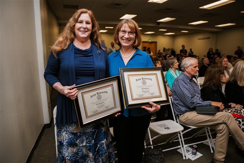 Two women smile while holding framed Avila University certificates at an award ceremony, with an audience seated in the background.